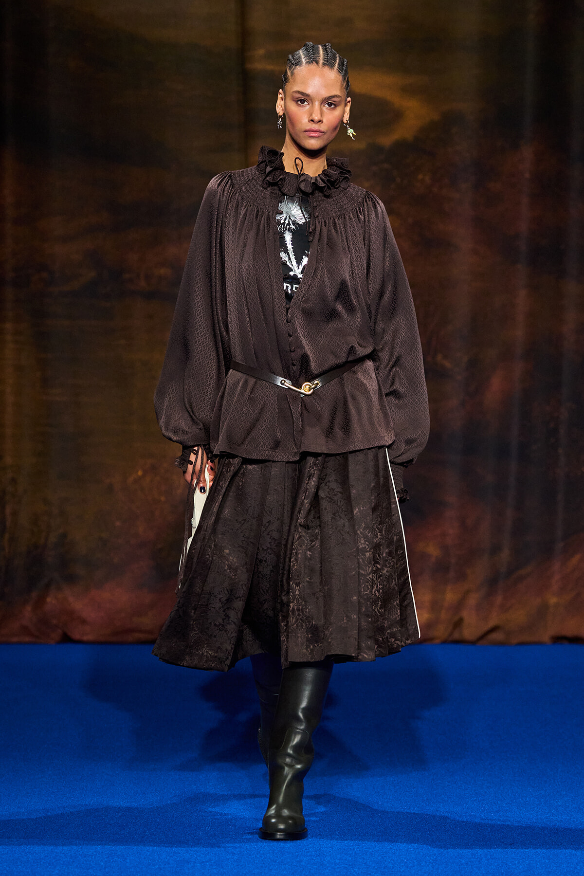 Runway model wearing a dark brown, textured blouse with a ruffled collar, belted waist, and a black lace skirt with knee-high boots on a blue catwalk surface.