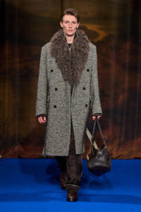Male model on a blue runway wearing a long gray tweed double-breasted coat with a large brown fur scarf and a black leather handbag.