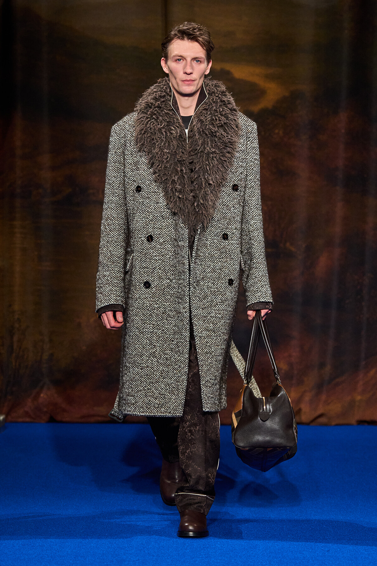 Male model on a blue runway wearing a long gray tweed double-breasted coat with a large brown fur scarf and a black leather handbag.