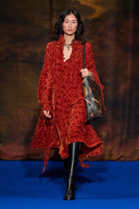 Model walking a fashion show in a red velvety, patterned dress with black knee-high boots and a large plaid shoulder bag on a blue runway.