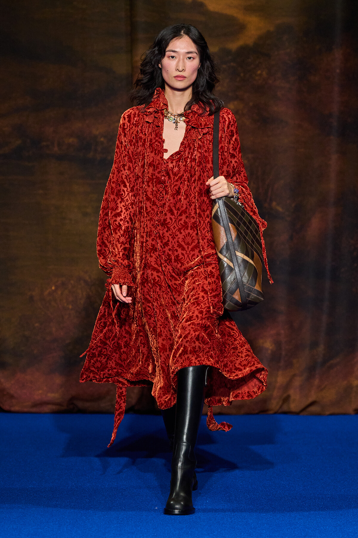Model walking a fashion show in a red velvety, patterned dress with black knee-high boots and a large plaid shoulder bag on a blue runway.
