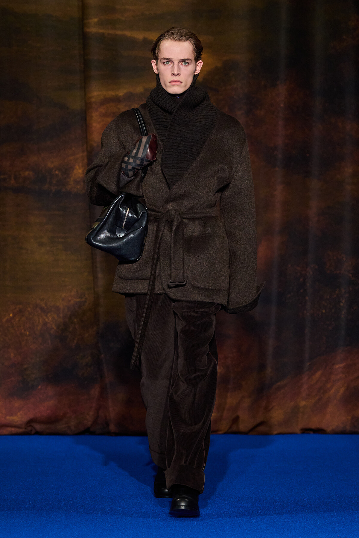 Male model walks a fashion runway in a dark brown belted wool coat and black turtleneck scarf, carrying a black bag over the shoulder.