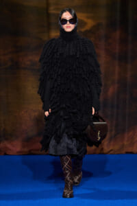 Model walking a fashion runway in a oversized black textured poncho with hood, sunglasses, and a brown handbag