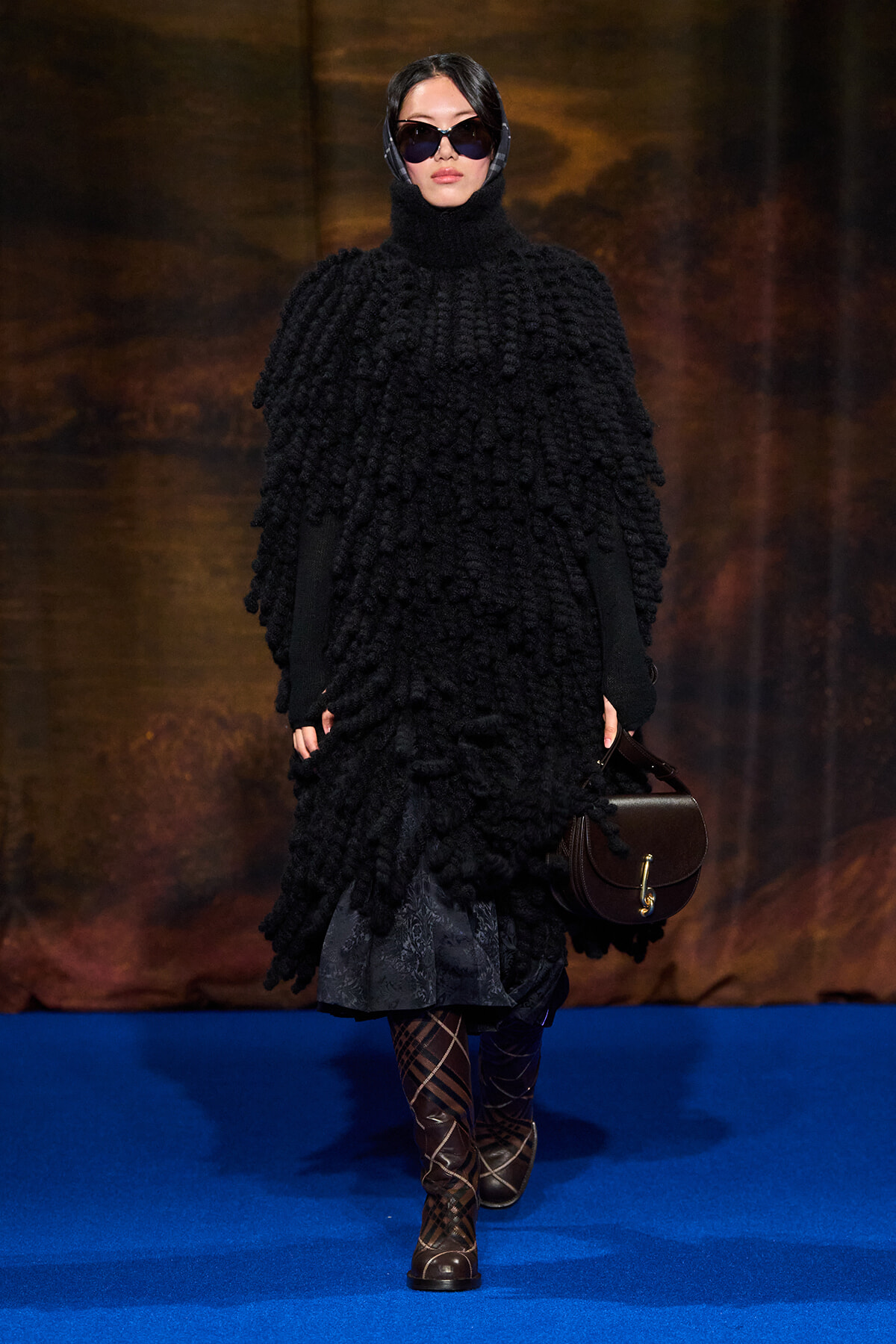 Model walking a fashion runway in a oversized black textured poncho with hood, sunglasses, and a brown handbag