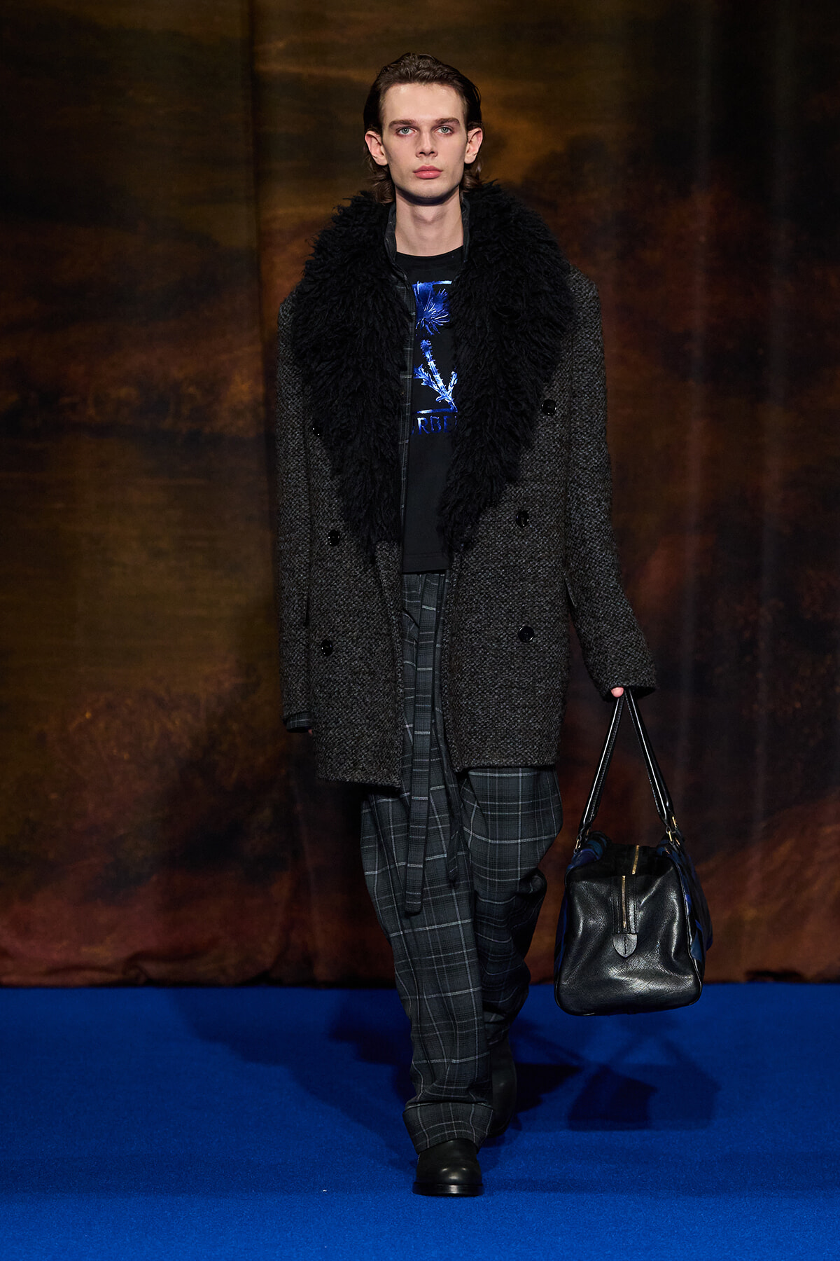 Male model on a runway wearing a textured dark coat with a shaggy black fur collar, plaid trousers, and a black leather handbag.