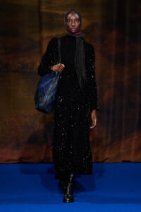 Model in a full-length black sequin dress and burgundy headscarf, carrying a blue plaid shoulder bag on a blue runway home to fashion show setting.