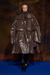 Model in an oversized brown shiny parka with a fur-trimmed hood walking a runway on a blue floor, boots visible