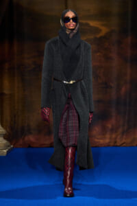 Model walks a blue runway in a long dark coat, heavy knit scarf, burgundy plaid skirt, red gloves and boots with sunglasses.