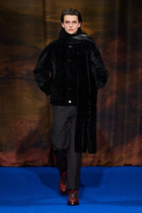 Model walking a runway in a oversized black fur coat, dark trousers, and red plaid boots on a blue catwalk backdrop