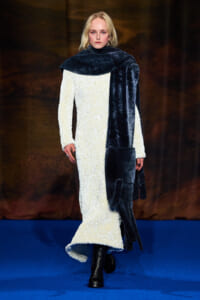 Model walking a fashion runway in a cream textured full-length dress with a dark fur scarf and black boots, against a blue runway backdrop.