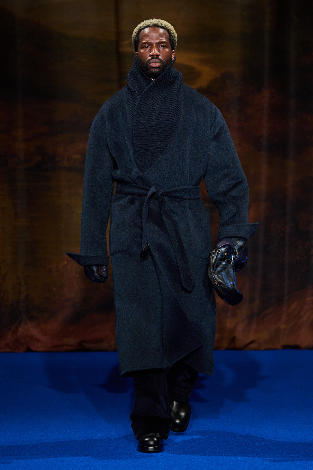 Male model on a blue runway wearing a navy wool wrap coat with belt, a thick matching scarf, gloves, and black boots, holding a plaid bag.