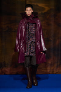 Model wearing a glossy burgundy parka over a patterned inner layer, dark pants, and brown knee-high boots on a blue runway stage.
