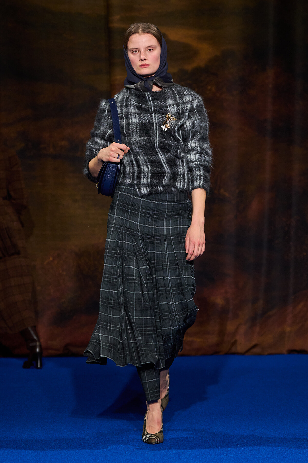 Runway model walking a fashion show in a gray plaid skirt and fuzzy black sweater, with a navy scarf and blue shoulder bag on a blue runway.