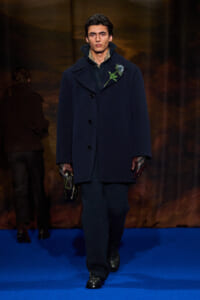 Male model walking a runway in a dark navy, oversized wool coat with a wide collar and hidden buttons, a blue flower boutonniere pinned at the chest; he wears fingerless gloves and dark knit pants, carrying a small bag in one hand against a blue runway.