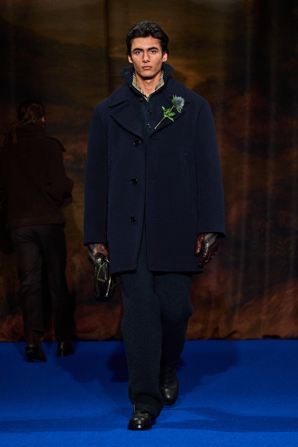 Male model walking a runway in a dark navy, oversized wool coat with a wide collar and hidden buttons, a blue flower boutonniere pinned at the chest; he wears fingerless gloves and dark knit pants, carrying a small bag in one hand against a blue runway.