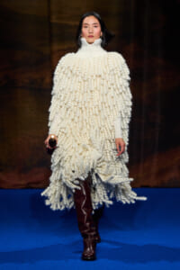 Model on a runway wearing a voluminous white chunky knit poncho with a turtleneck and brown knee-high boots.