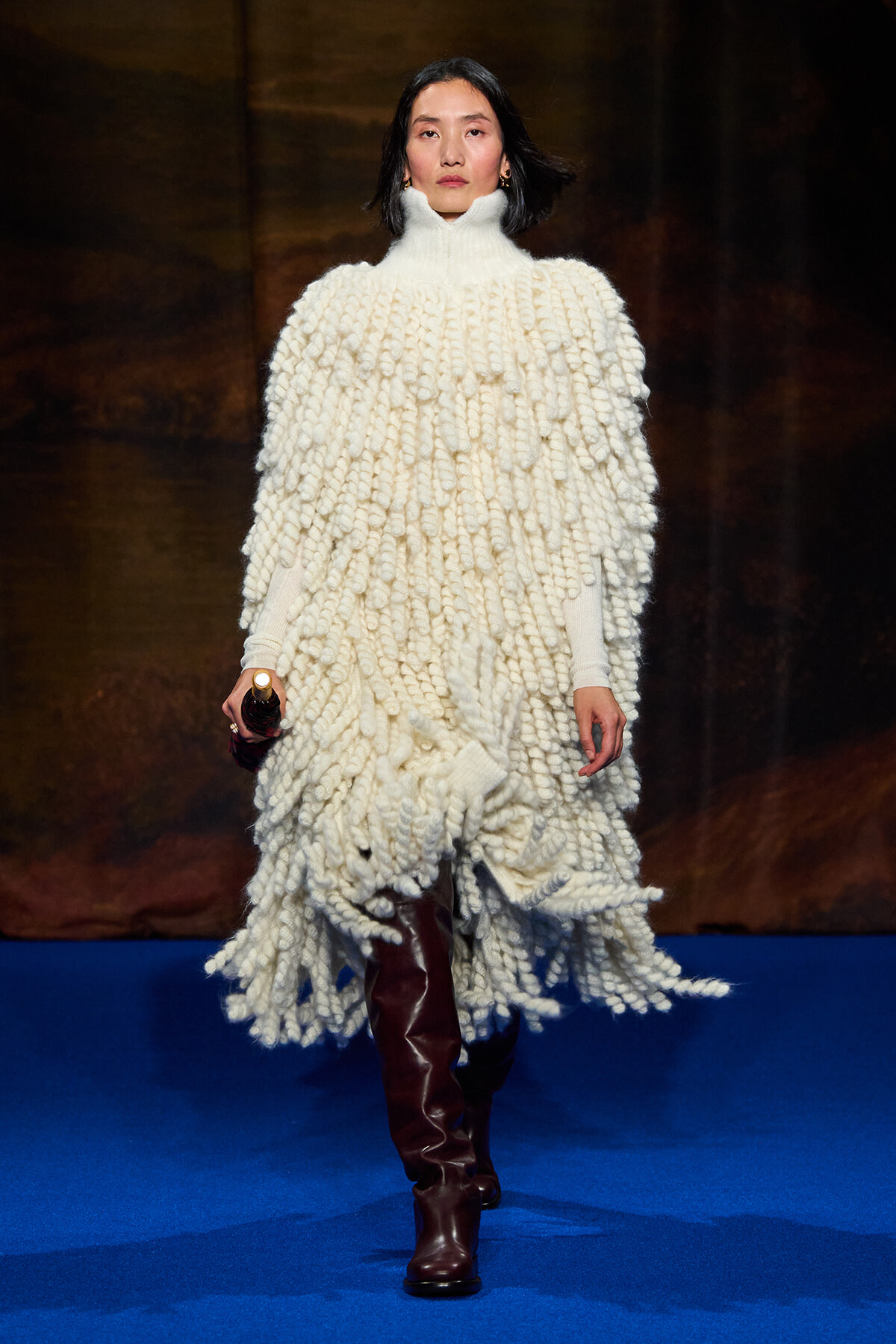 Model on a runway wearing a voluminous white chunky knit poncho with a turtleneck and brown knee-high boots.