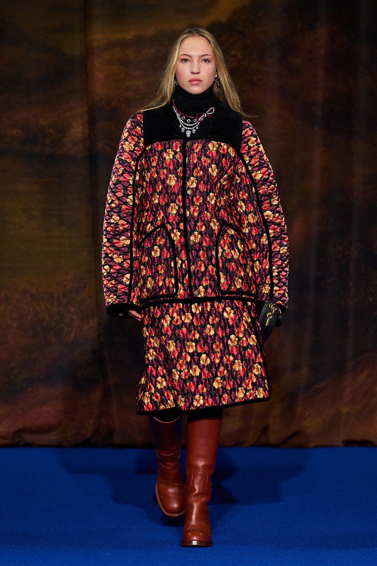 Model walks the runway in a red-yellow floral quilted coat with black trim and brown knee-high boots.