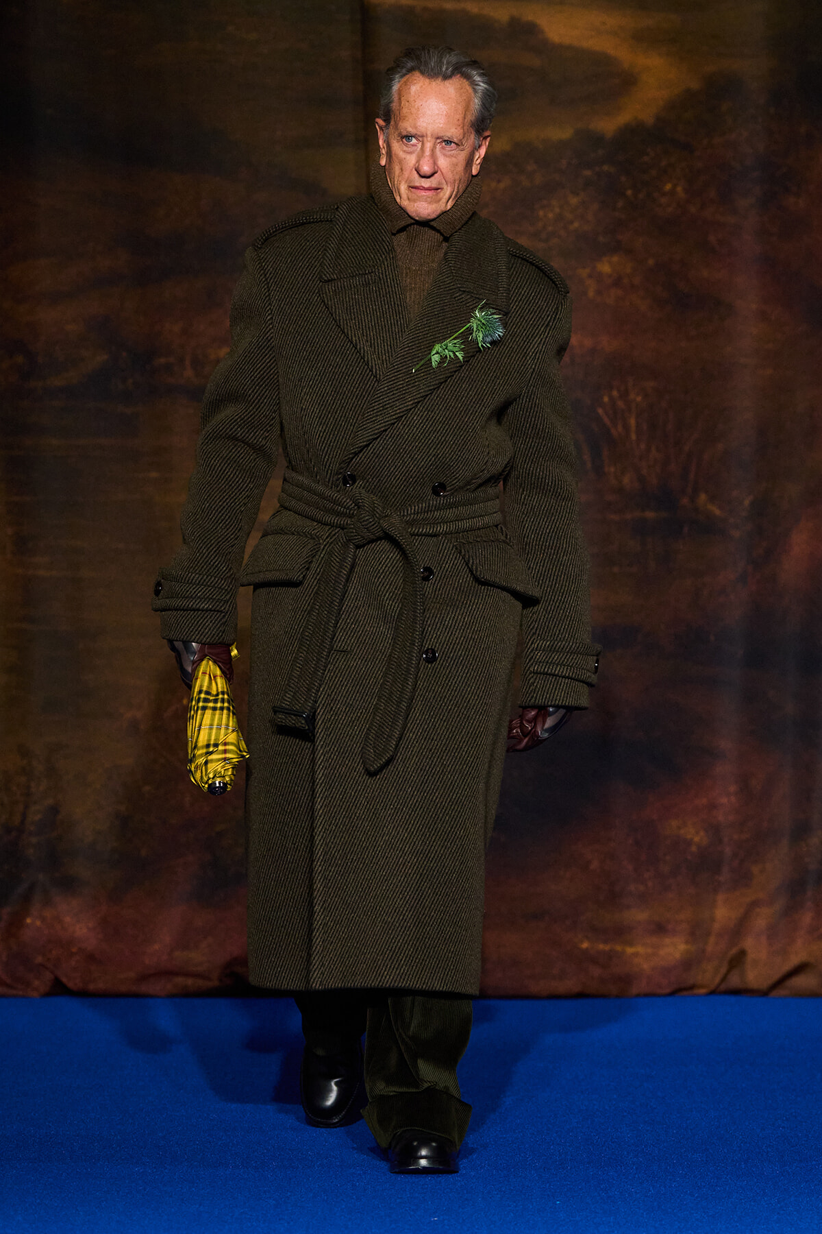 Man walking a fashion show in a long olive wool coat with a belt, dark gloves, and a yellow plaid umbrella on a blue runway.