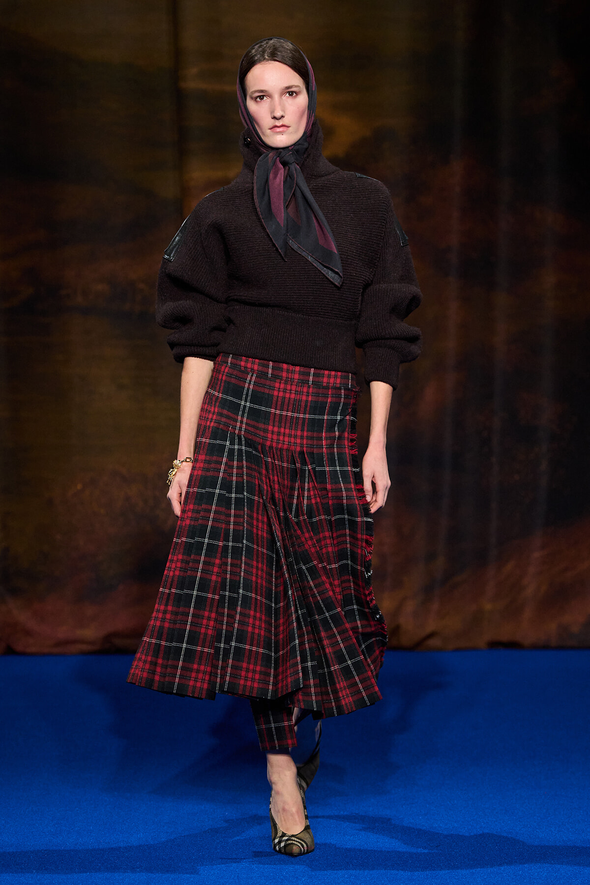 Model on a blue runway wearing a dark oversized knit sweater, a red plaid midi skirt, and a scarf tied at the neck; walking forward on the catwalk.