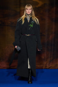 Model walking a fashion show in a long black wool coat with a plaid scarf, gold belt, and a green floral pin, clutch in hand against a blue runway.