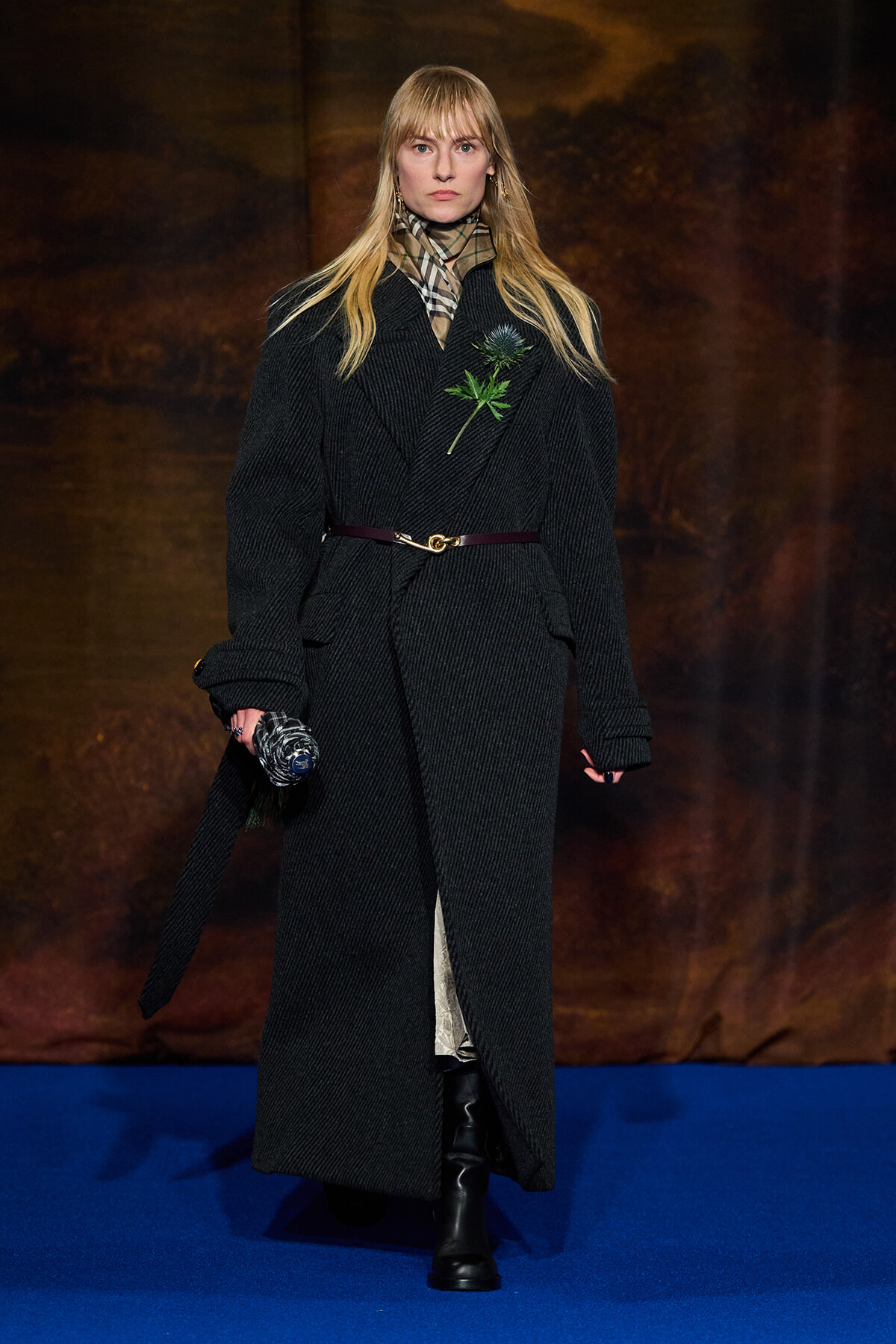 Model walking a fashion show in a long black wool coat with a plaid scarf, gold belt, and a green floral pin, clutch in hand against a blue runway.