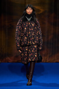 Model on a blue runway wearing a black floral quilted coat, black turtleneck, layered necklaces, and brown knee-high boots.