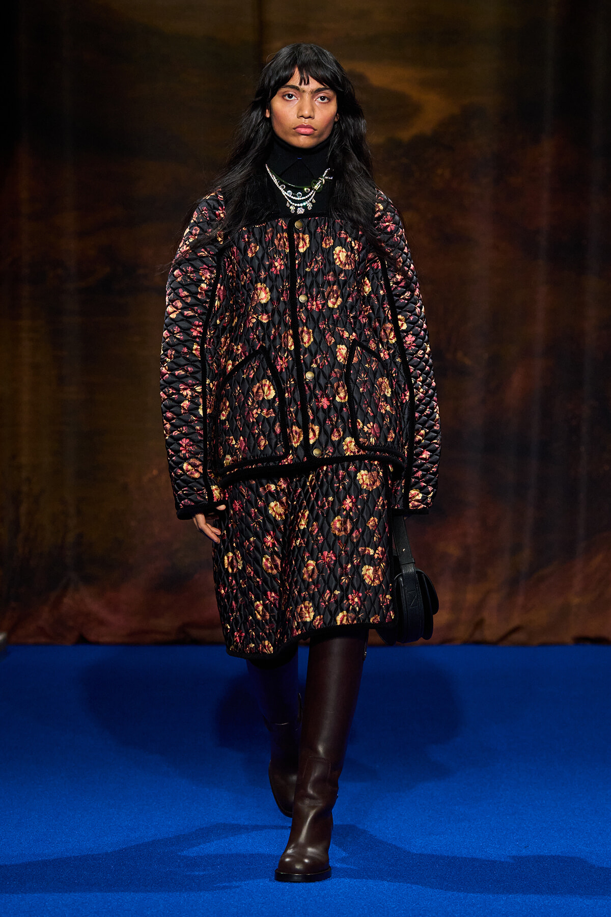 Model on a blue runway wearing a black floral quilted coat, black turtleneck, layered necklaces, and brown knee-high boots.