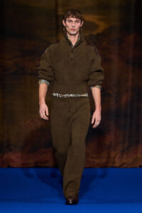Male model walks a runway wearing a brown knit pullover with shoulder patches and matching knit pants, plaid shirt peeking at the cuffs from underneath.