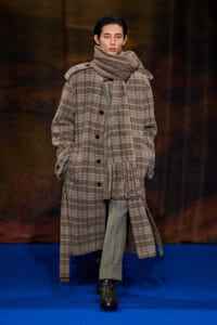 Model walking a runway in a oversized brown plaid coat with a chunky scarf and green trousers on a blue catwalk