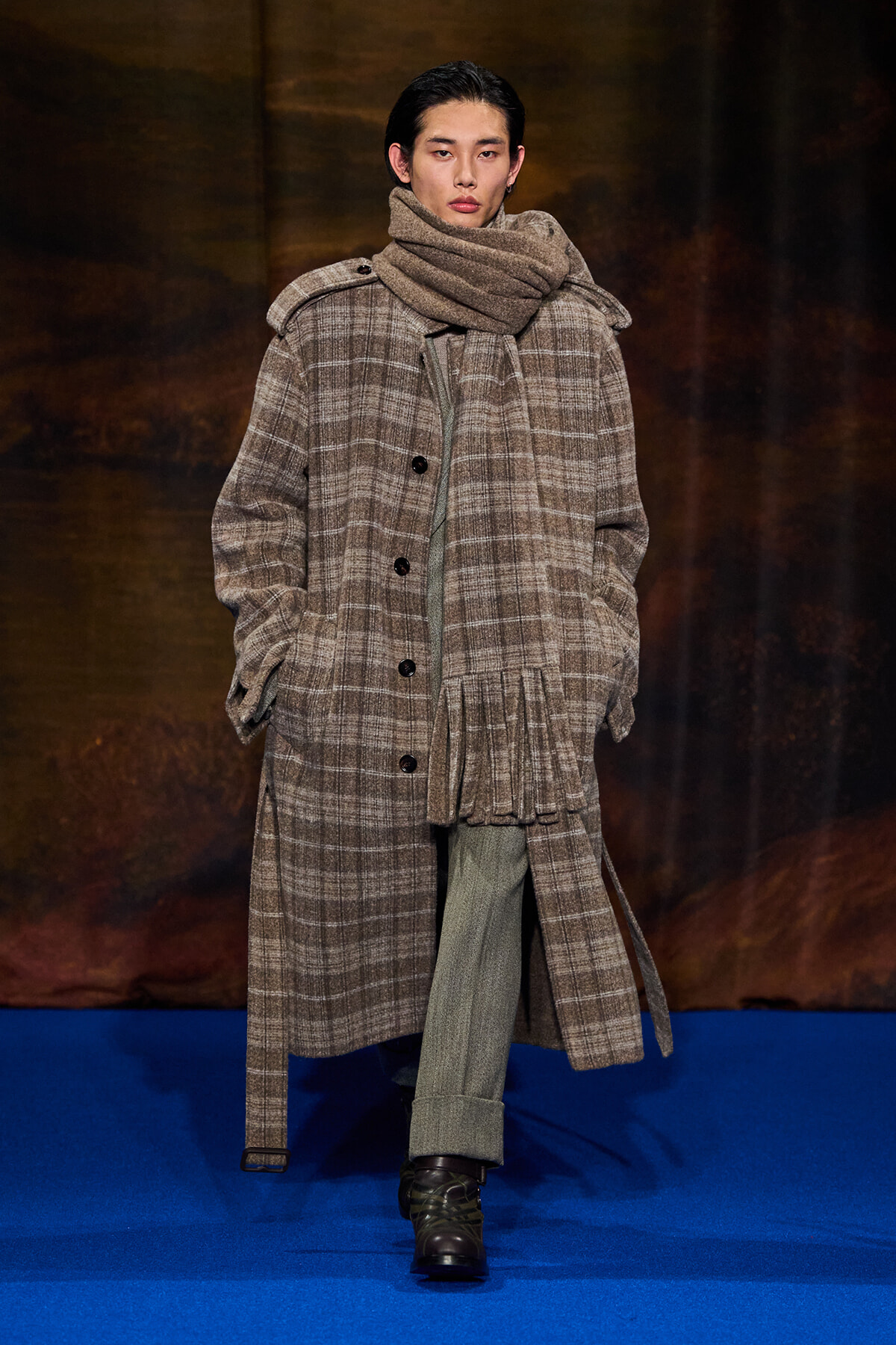 Model walking a runway in a oversized brown plaid coat with a chunky scarf and green trousers on a blue catwalk