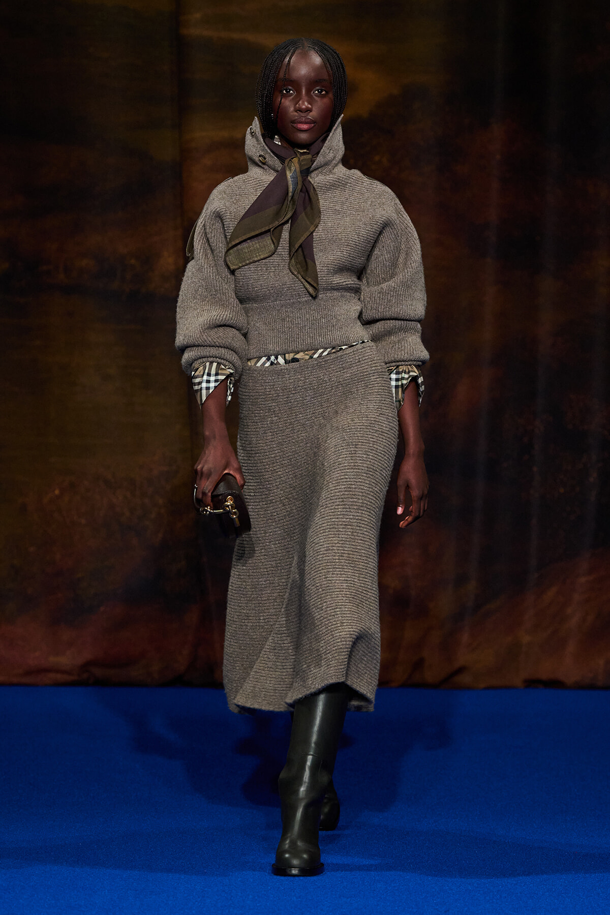 Model walking a runway in a taupe knit sweater and matching skirt, with a scarf tied at the neck and plaid cuffs showing under the sleeves, holding a small clutch.