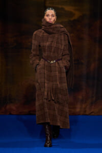 Model walking the runway in a brown plaid wool coat with a large scarf draped around the neck and a purple belt.