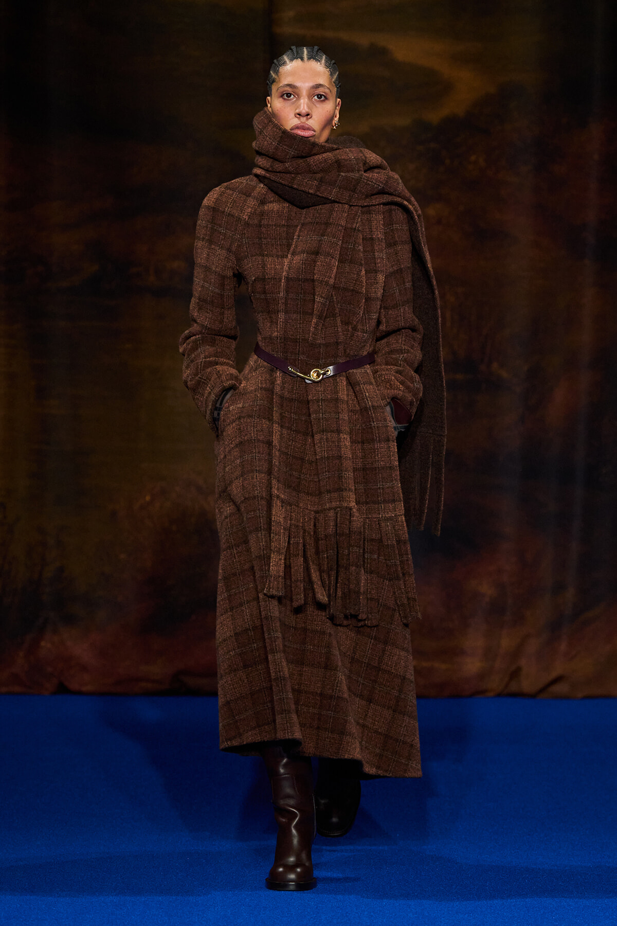 Model walking the runway in a brown plaid wool coat with a large scarf draped around the neck and a purple belt.