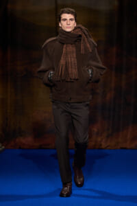 Male model walking runway in a brown wool coat with matching scarf and boots, hands in pockets on a blue catwalk.