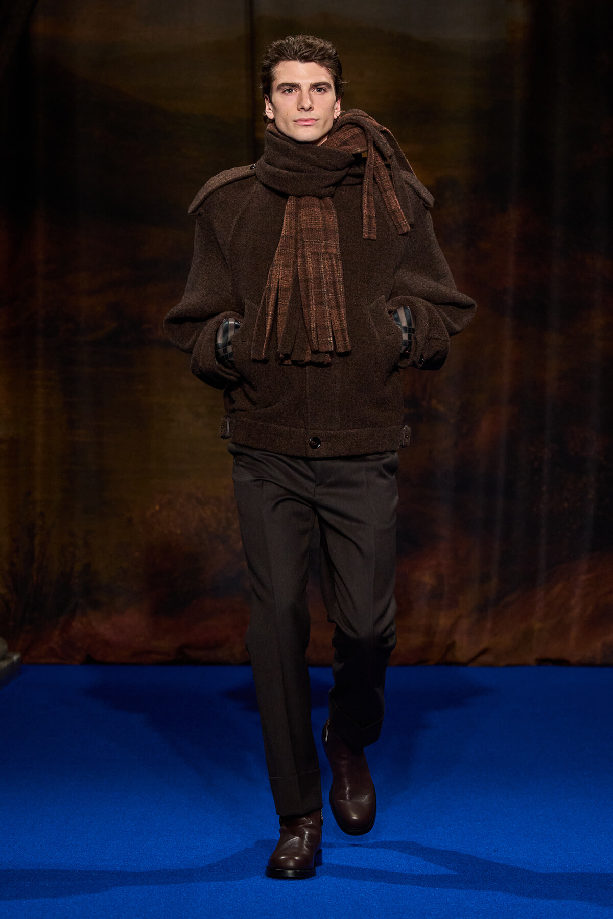 Male model walking runway in a brown wool coat with matching scarf and boots, hands in pockets on a blue catwalk.