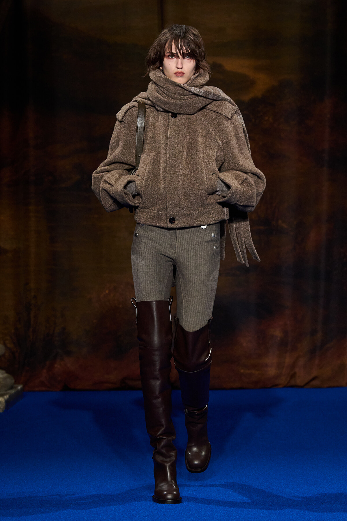 Model walking the runway in a bulky taupe-brown coat with a large scarf, gray knit pants, and tall brown knee-high boots on a blue catwalk background