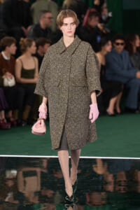 Model walking a runway in a speckled oversized coat, pale pink gloves, and a small pink handbag, against a green floor and backstage audience.