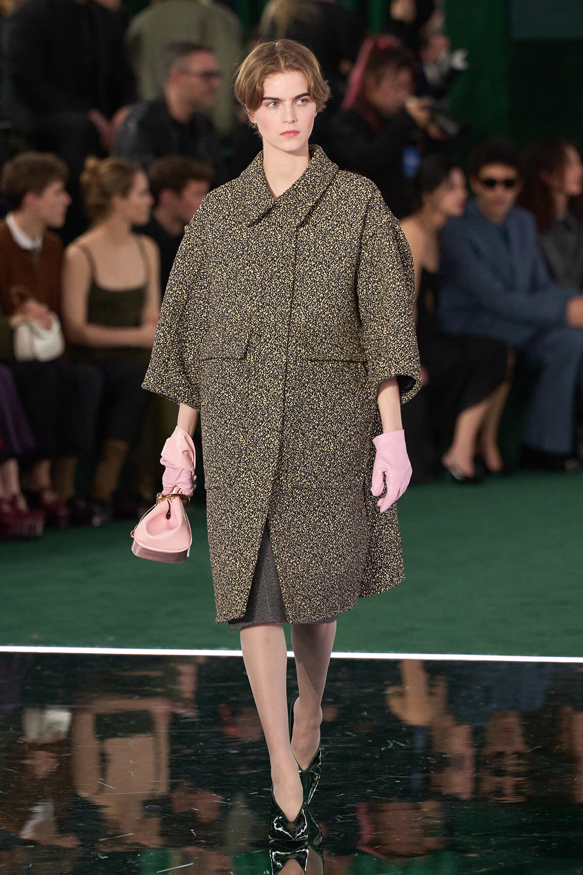 Model walking a runway in a speckled oversized coat, pale pink gloves, and a small pink handbag, against a green floor and backstage audience.