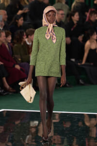 Model walking the runway in a green textured suit with a tied peach scarf and beige hat, carrying a cream handbag.
