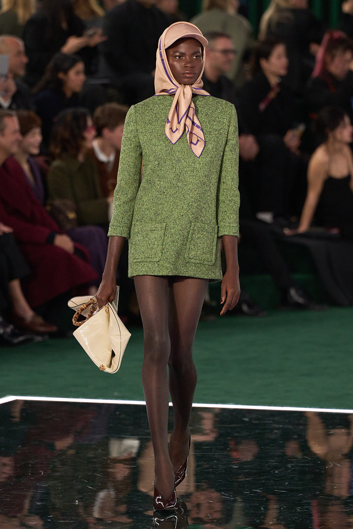 Model walking the runway in a green textured suit with a tied peach scarf and beige hat, carrying a cream handbag.