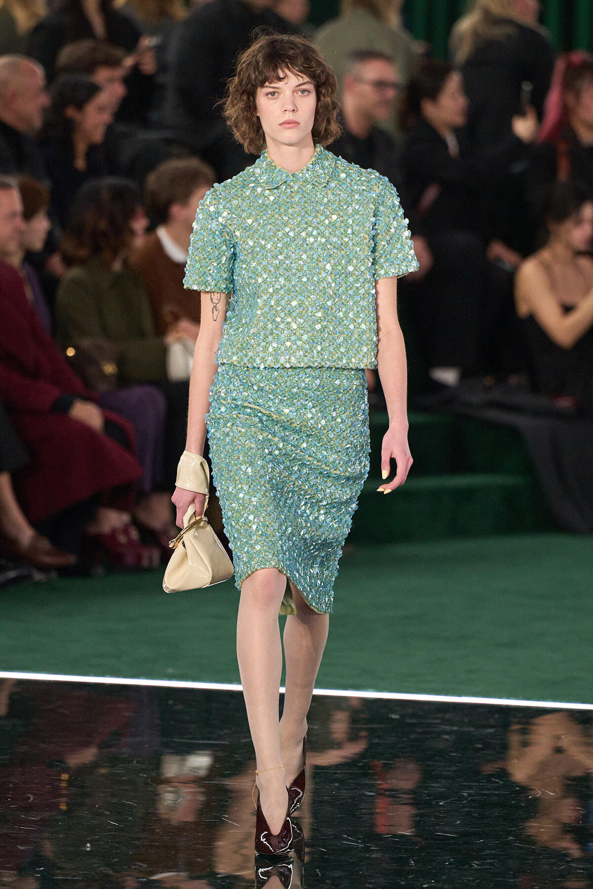 Model walks the runway in a seafoam green sequined two-piece outfit (short-sleeve top and knee-length skirt) with a beige handbag.