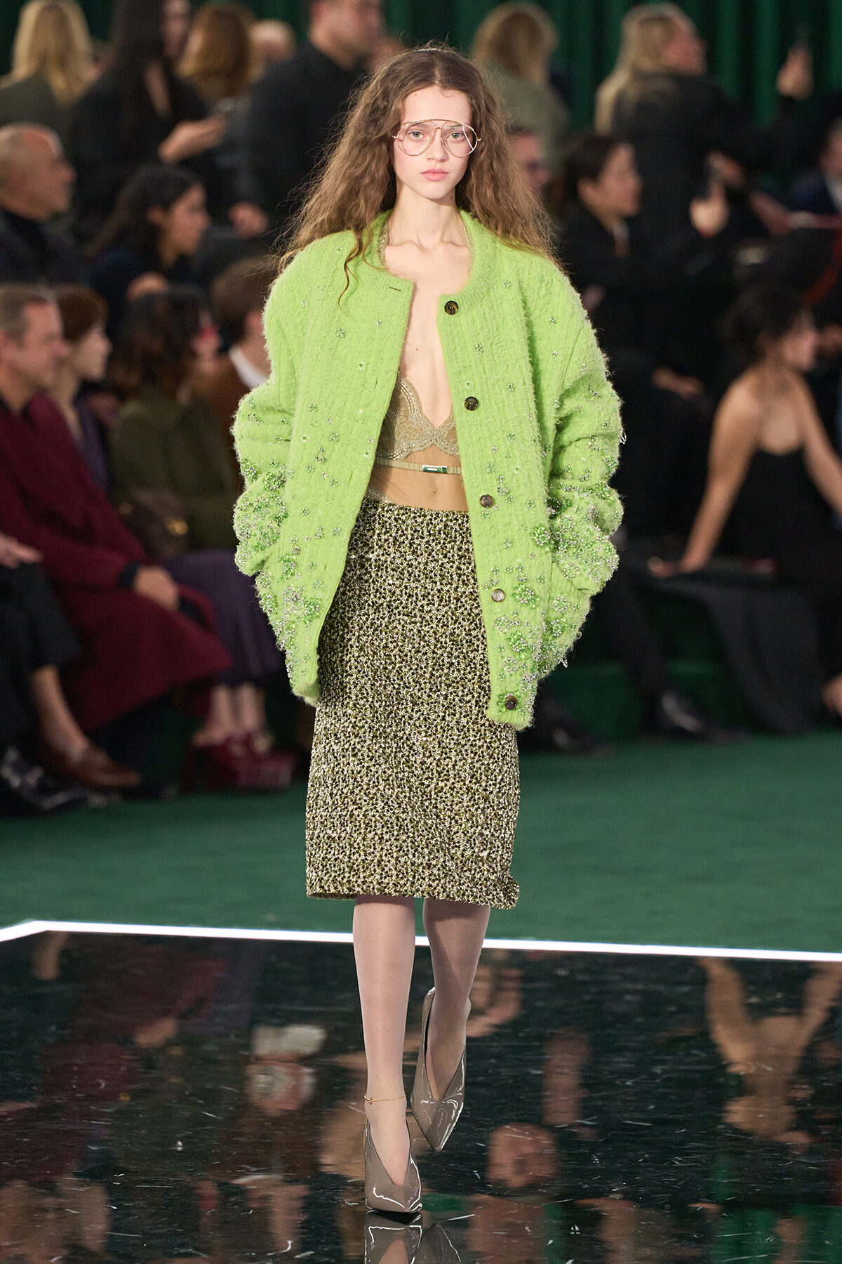 Runway model wearing a bright lime-green textured jacket over a leopard-print midi skirt with beige bralette and oversized glasses.
