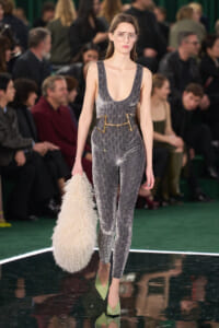 Model walks the runway in a metallic grey, patterned jumpsuit with a gold belt, lime-green heels, and oversized clear glasses, carrying a fluffy cream bag.
