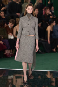 Model walks a gray tweed two-piece suit (cropped jacket and midi skirt) on a runway, holding a pink handbag and wearing burgundy heels.