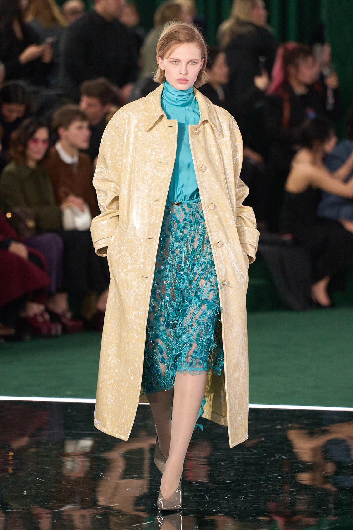 Model walks a runway in a light cream textured overcoat over a turquoise satin turtleneck and teal lace dress, with an audience in the background.