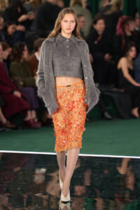 Model on a runway wearing a gray cropped knit sweater and an orange floral lace midi skirt, beige heels.