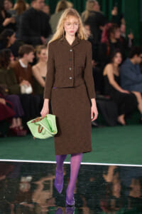 Runway model in a brown textured blazer and knee-length skirt, wearing glasses, carrying a light green handbag, and purple tights with matching purple heels.
