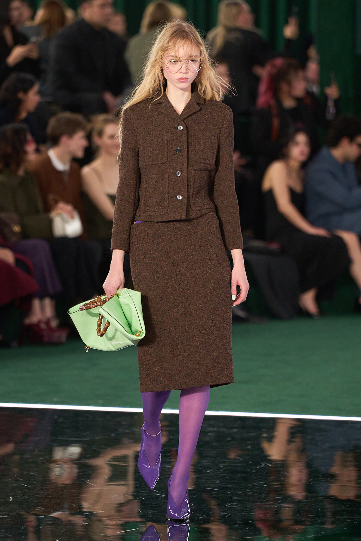 Runway model in a brown textured blazer and knee-length skirt, wearing glasses, carrying a light green handbag, and purple tights with matching purple heels.