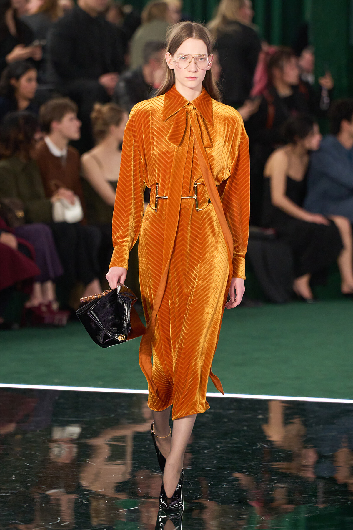 Model walks runway in velvety orange dress with tie-front neck, oversized belt, and large glasses; carries black patent bag and wears black heels.